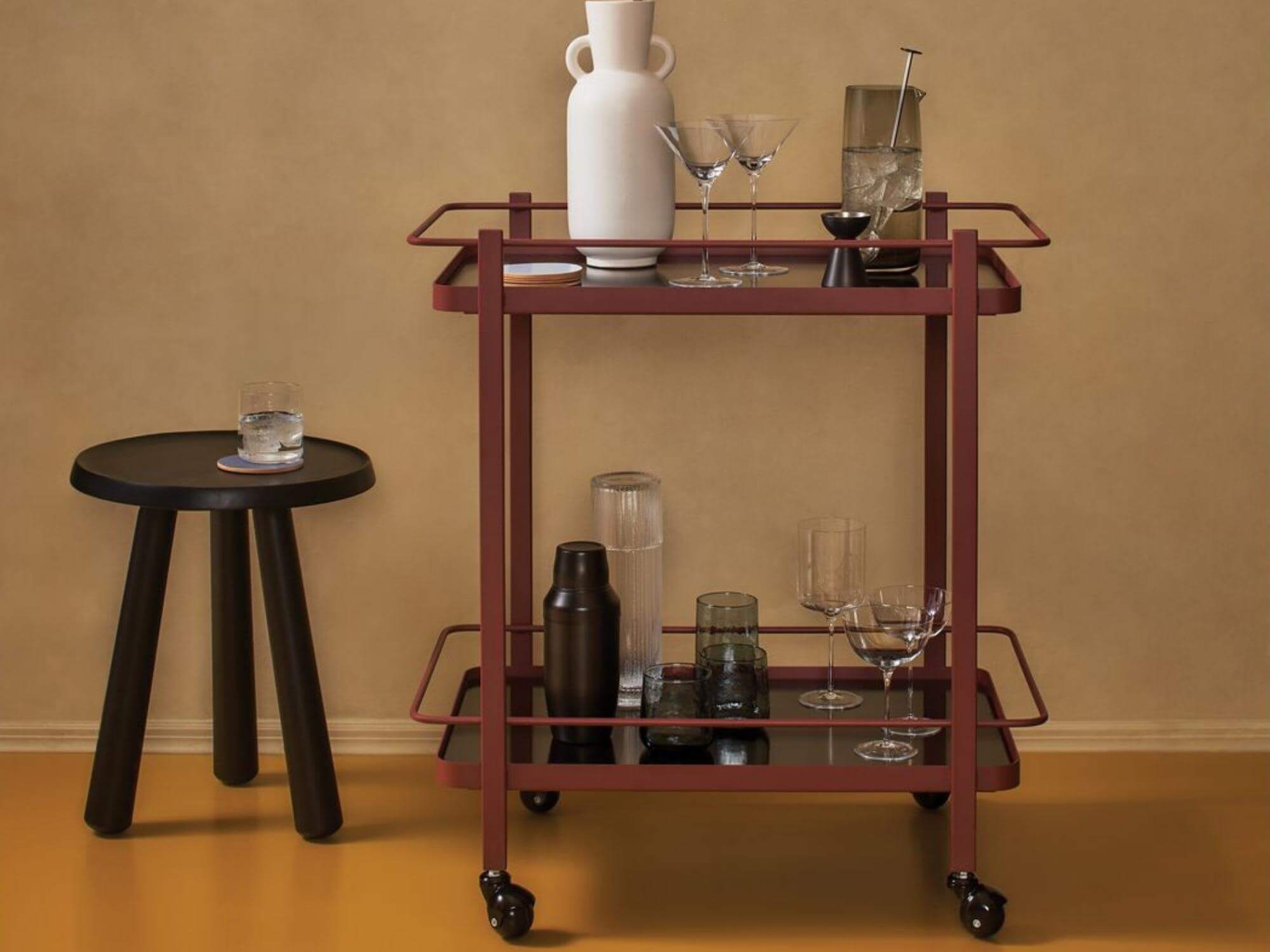 Black milk stool with glass of water on it beside red bar cart