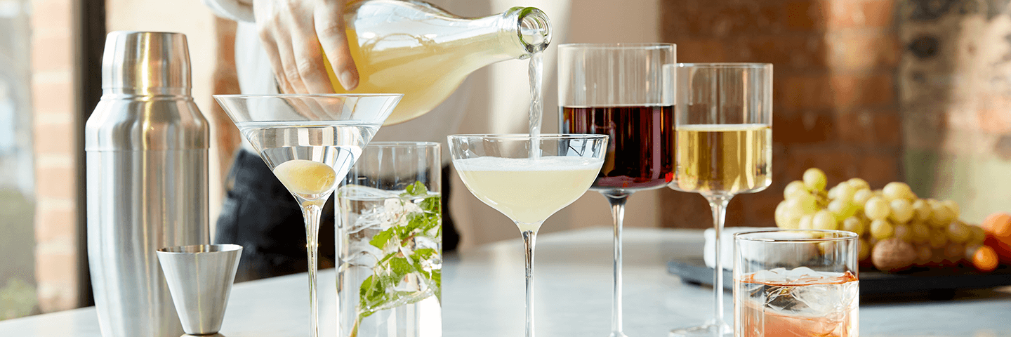 Hand pours pale liquid into coupe glass beside wine and cocktail glasses