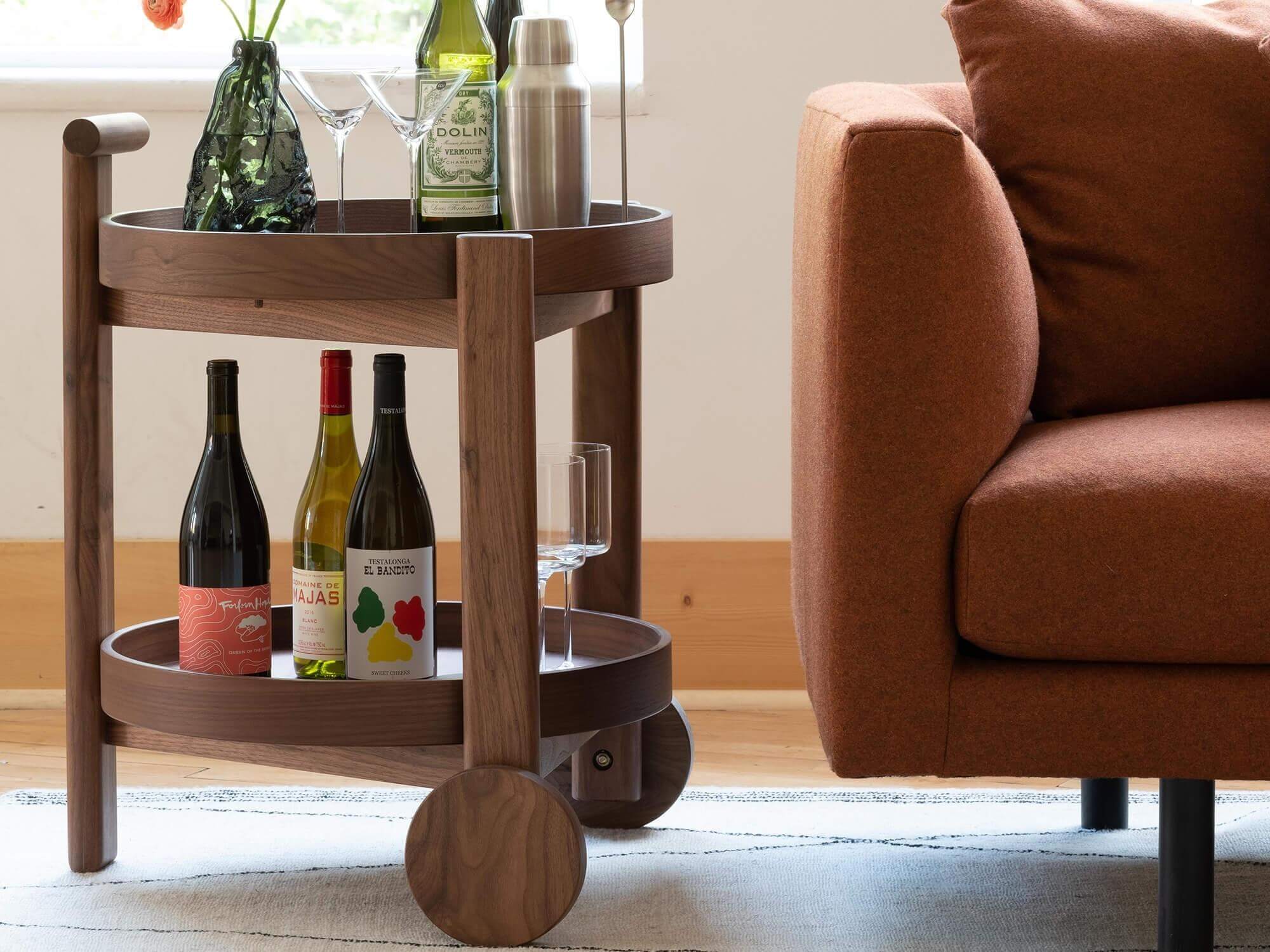 Two Vesper unique martini glasses sit on a crowded walnut bar cart beside a red sofa