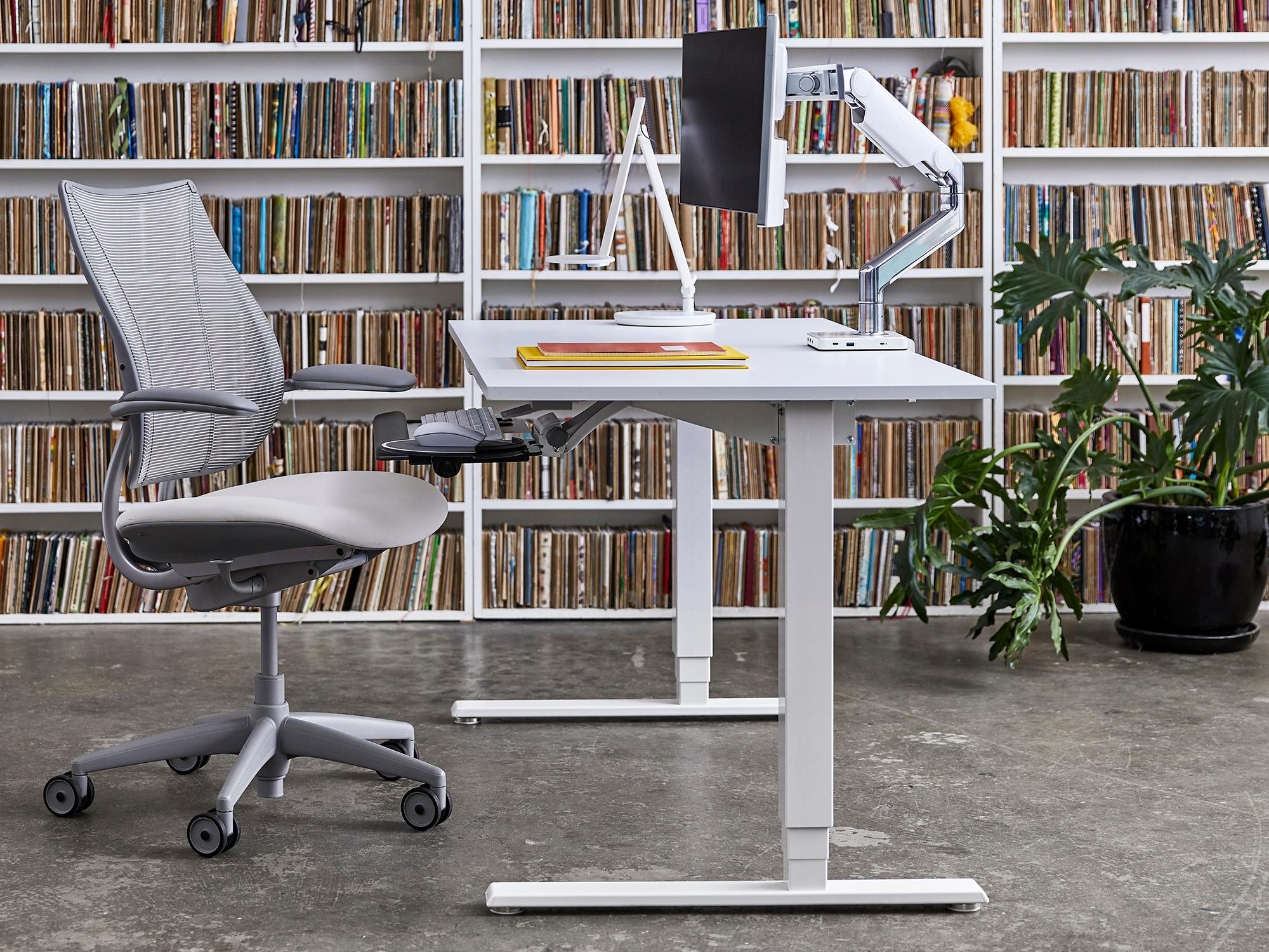 The Humanscale M8.1 Monitor Arm is set up on a white desk over a keyboard tray with mouse tray in front of a wall of books