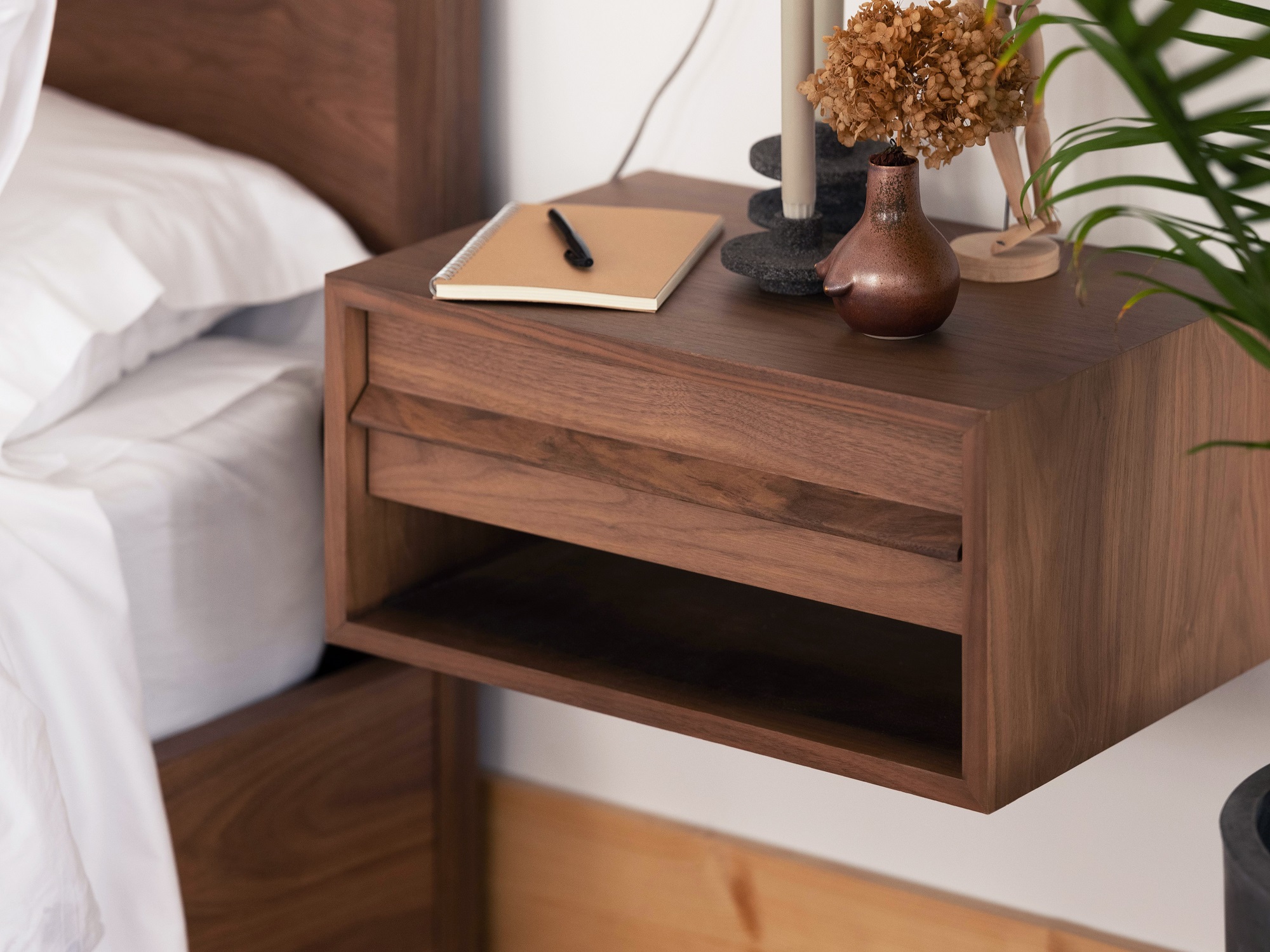 The walnut Marcel open shelf modern floating nightstand beside a bed is stacked with notebook, pen, and candlesticks