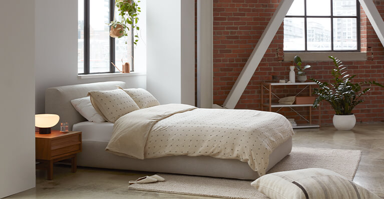 Queen size grey fabric bed is shown in a studio apartment setting. Link to beds category