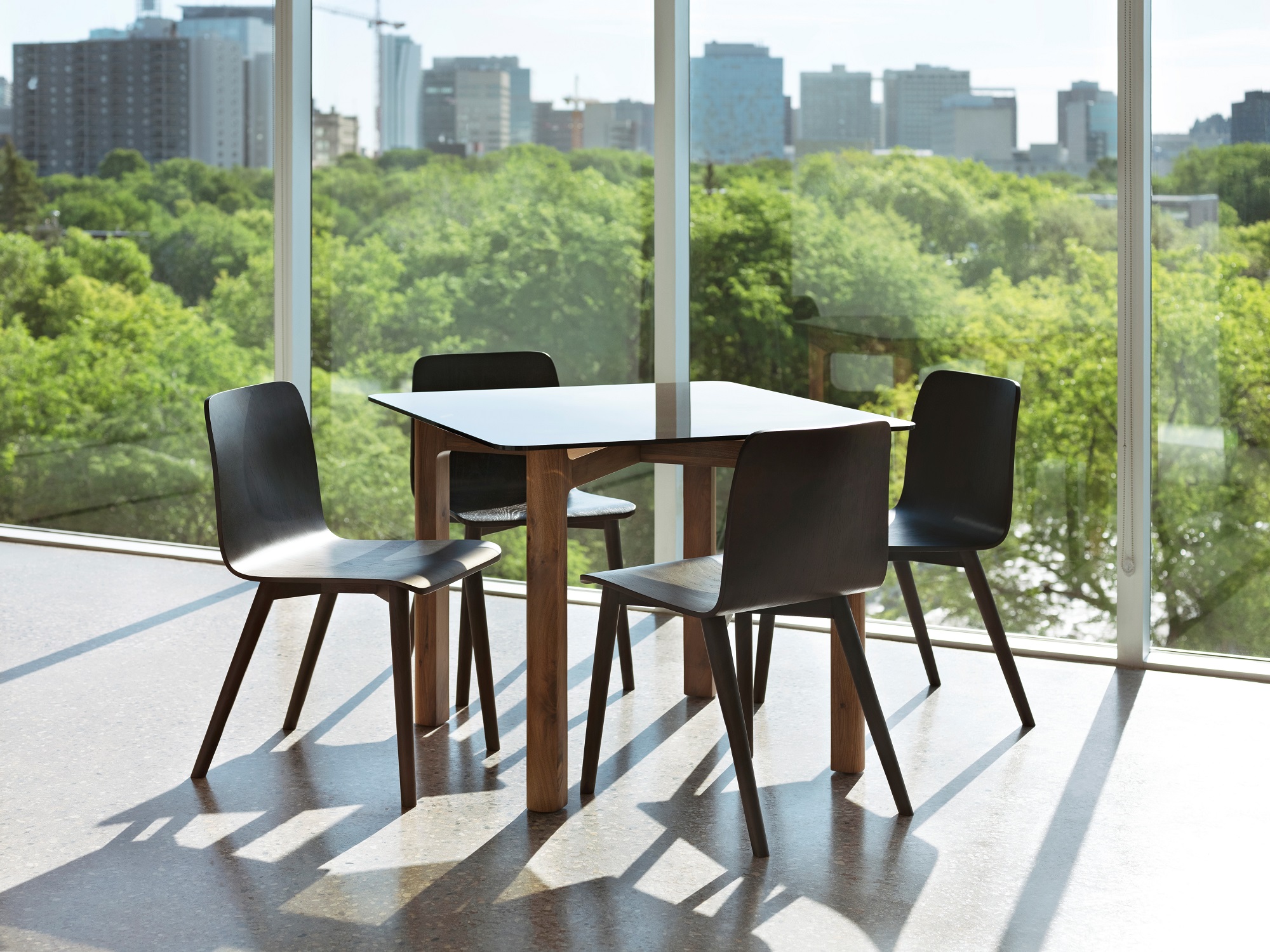 Grey glass top dinette table with walnut legs surrounded by black dining chairs in bright dining space