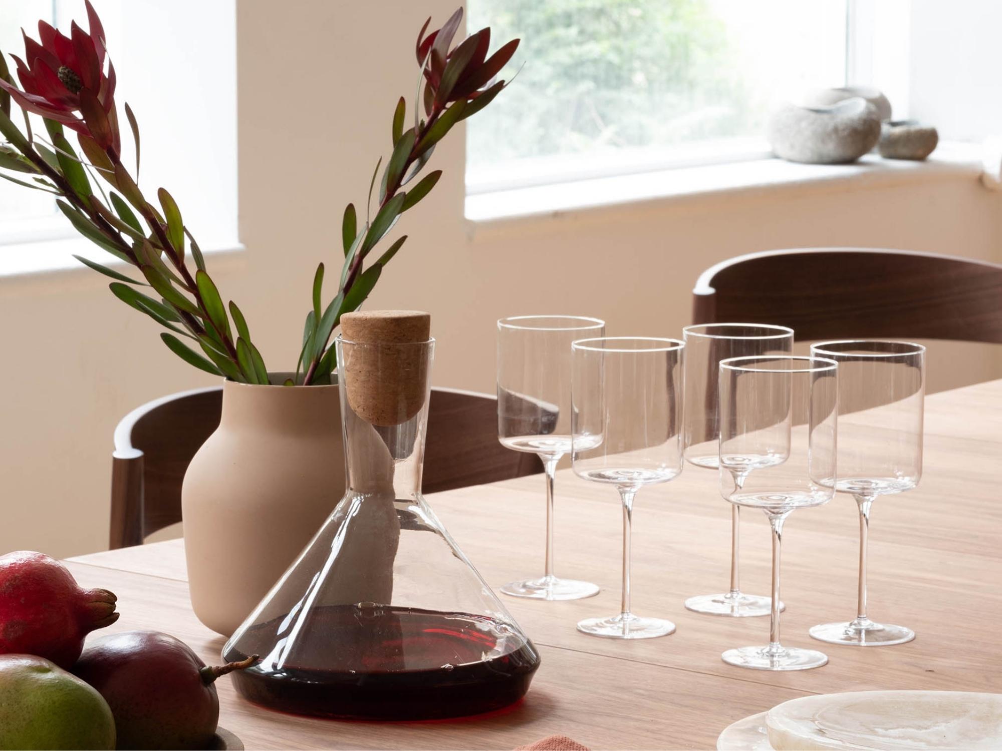 Six Vesper modern white wine glasses sit on a walnut dining table next to a decanter and ceramic vase with red flowers in it