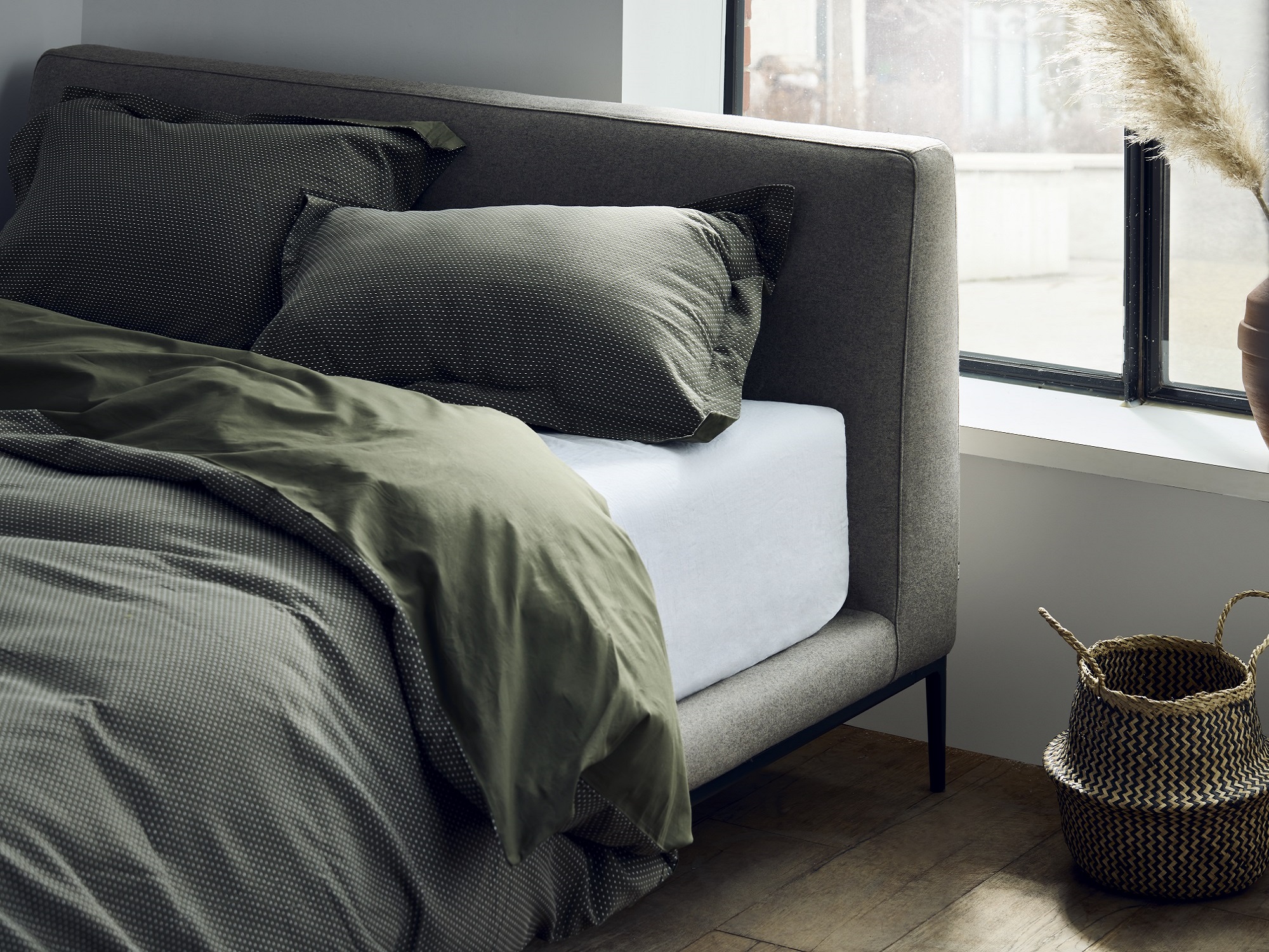 Corner of a grey upholstered bed with green bedding by a large bright window