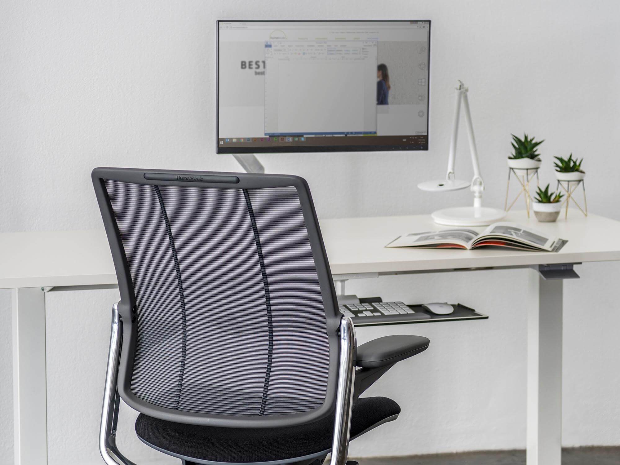 The Humanscale M2.1 Monitor Arm is set up on a white desk with an office chair in front of it