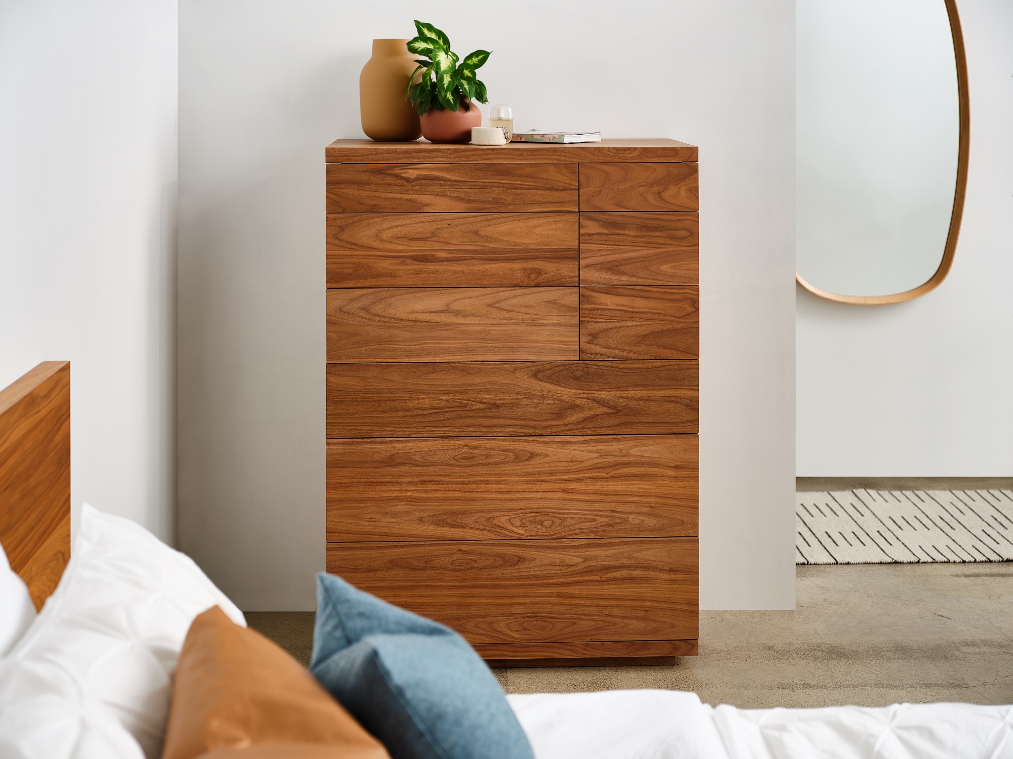 A tall walnut chest of drawers with vases on it sits in a white bedroom