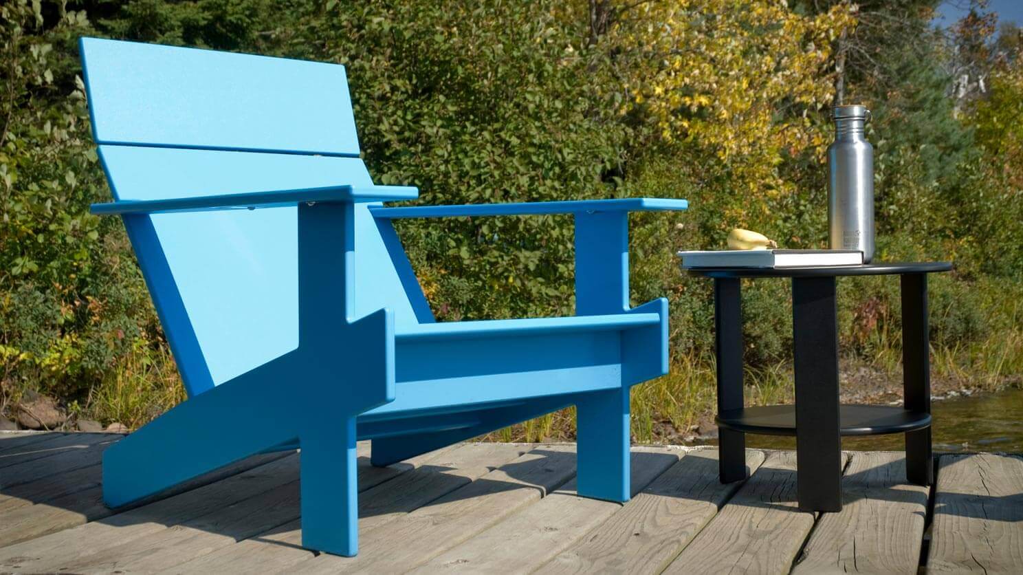 The blue Lollygagger patio lounge chair sits on a wooden deck with a forest behind it and an outdoor end table beside