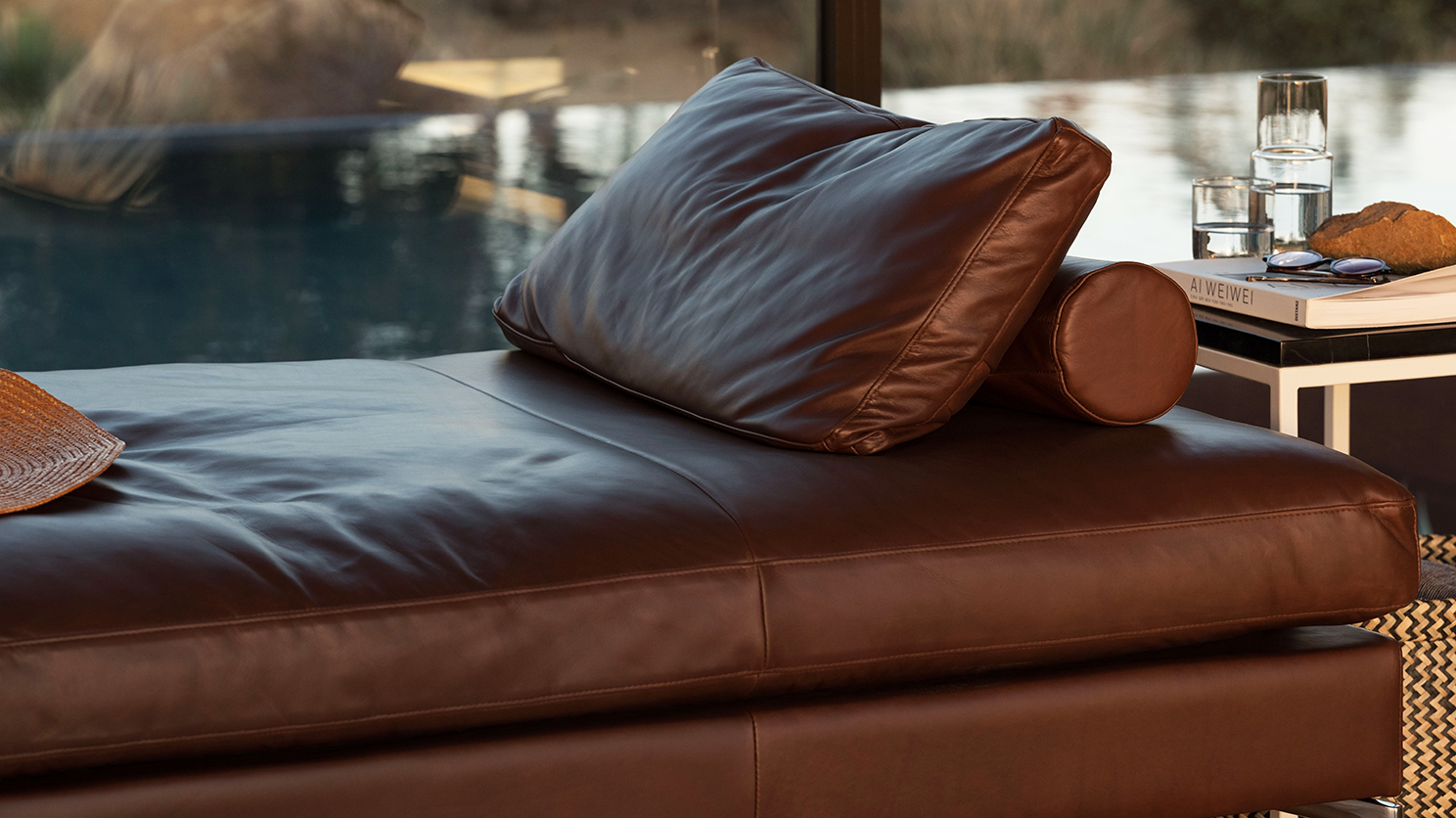 The Eve moern chaise lounge in brown leather in front of patio windows with a white end table beside it