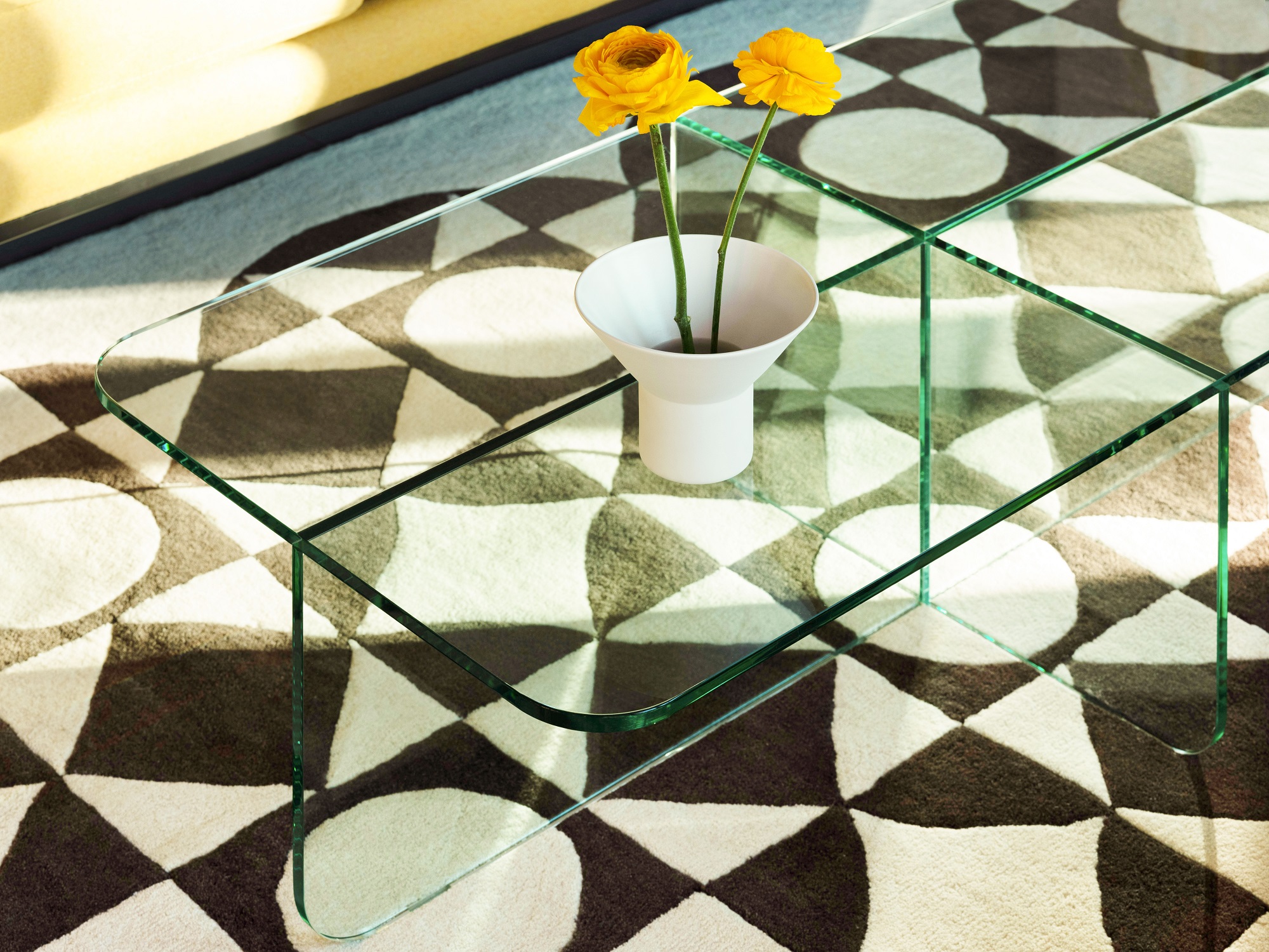 White vase with yellow flowers in it on glass rectangular coffee table with monochromatic geometric pattern rug