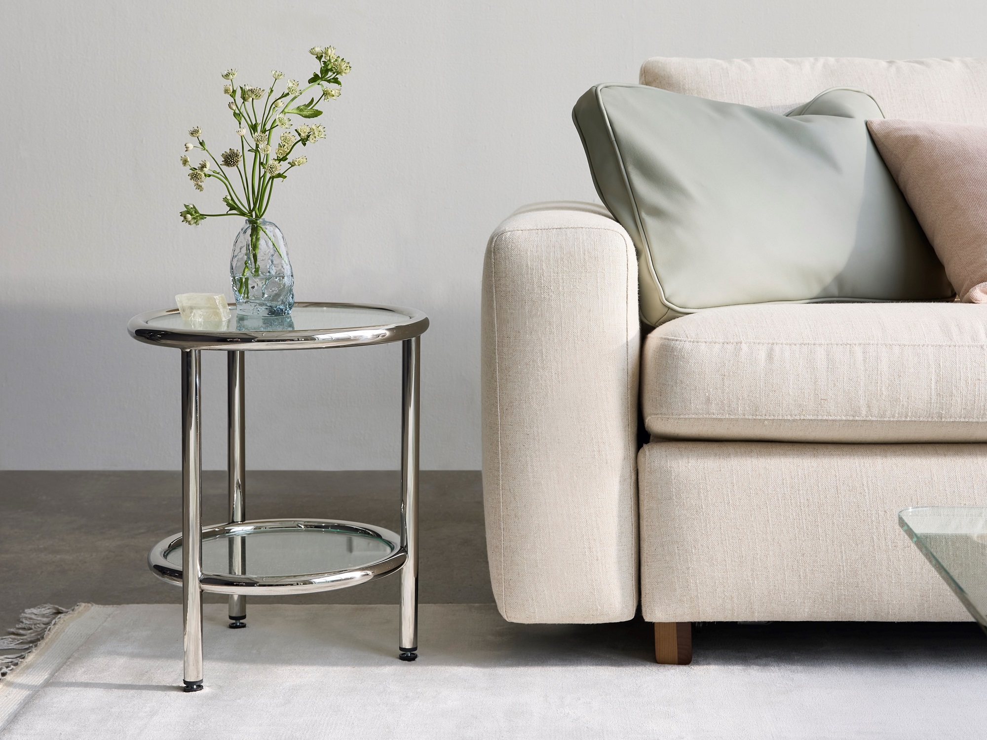 Stainless steel and glass end table with glass flower vase beside beige couch