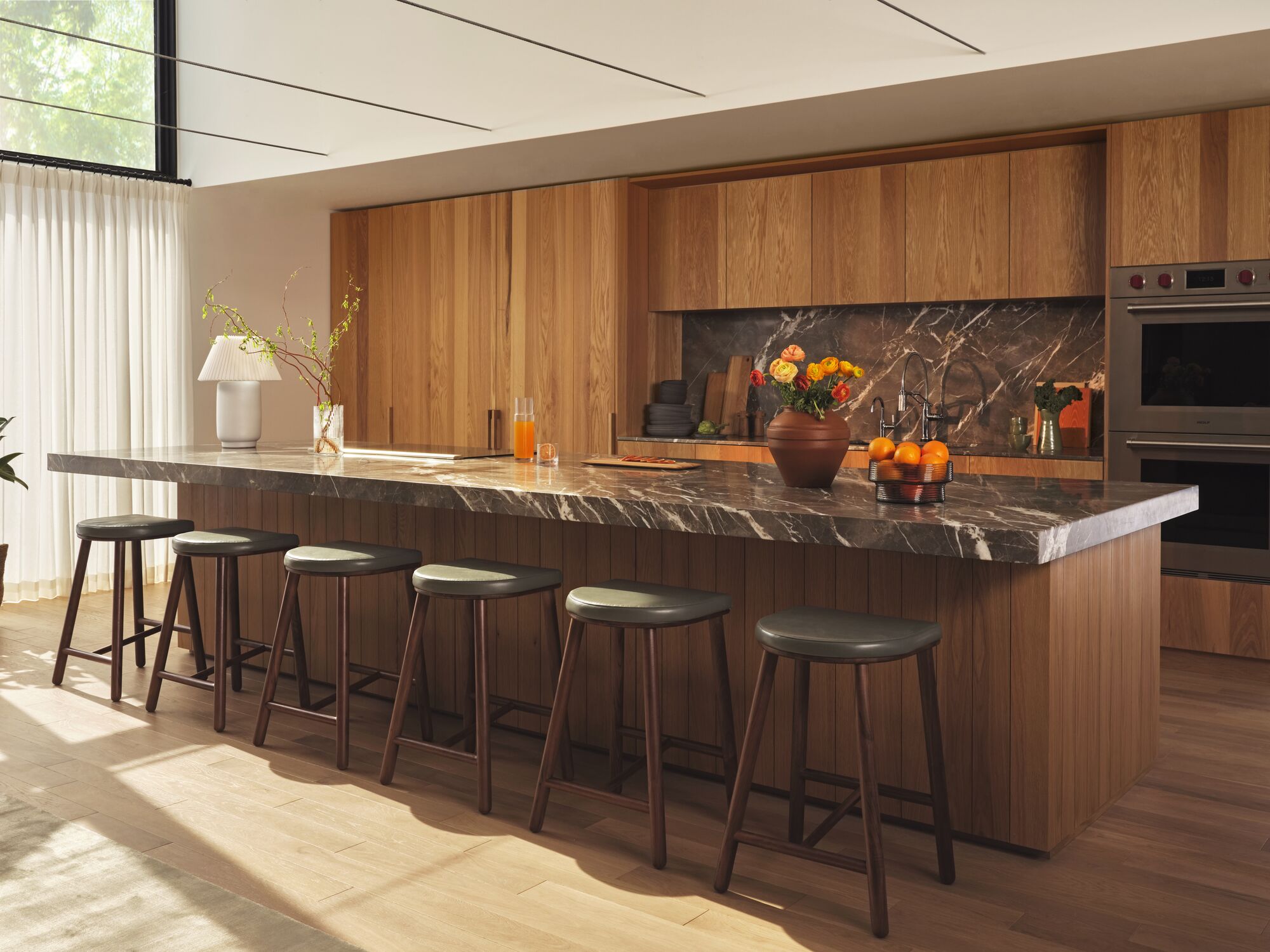 Grey leather and walnut stools lined along black marble counter in wood-paneled kitchen