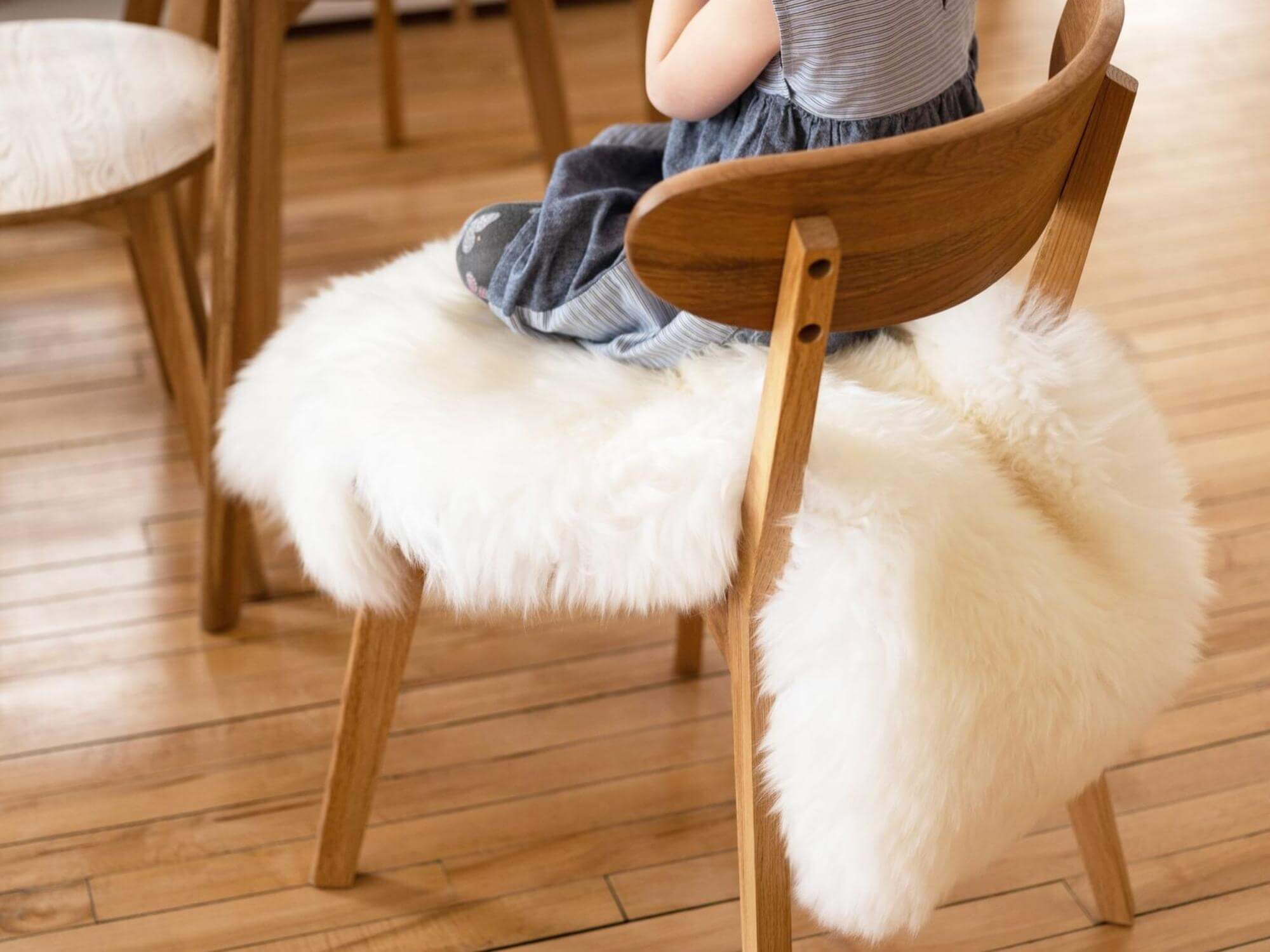 Little girl sits on a sheepskin rug across a dining chair pulled up to matching wood table