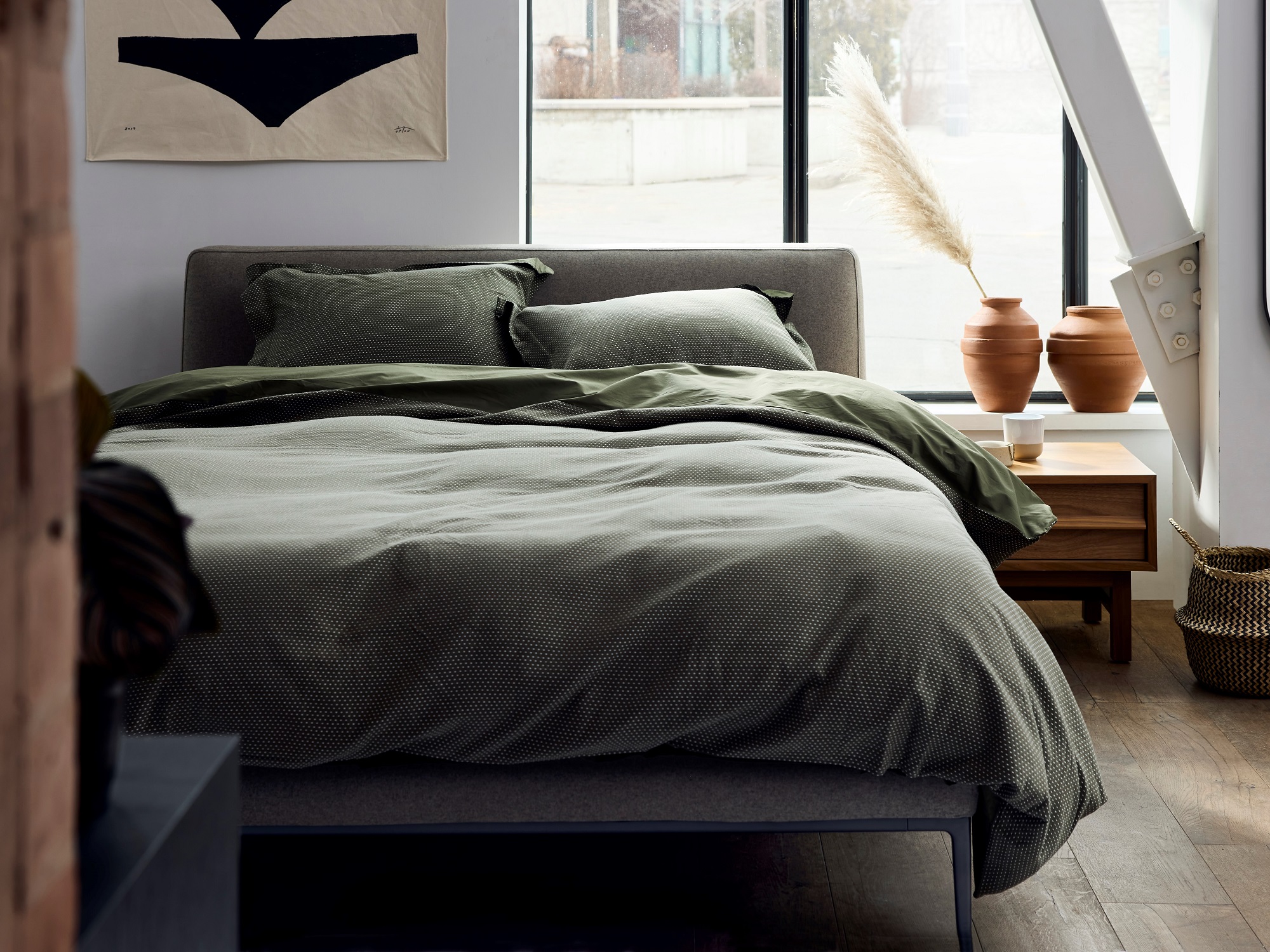 A gray upholstered bed with green bedding on it, in a white bedroom with modern art